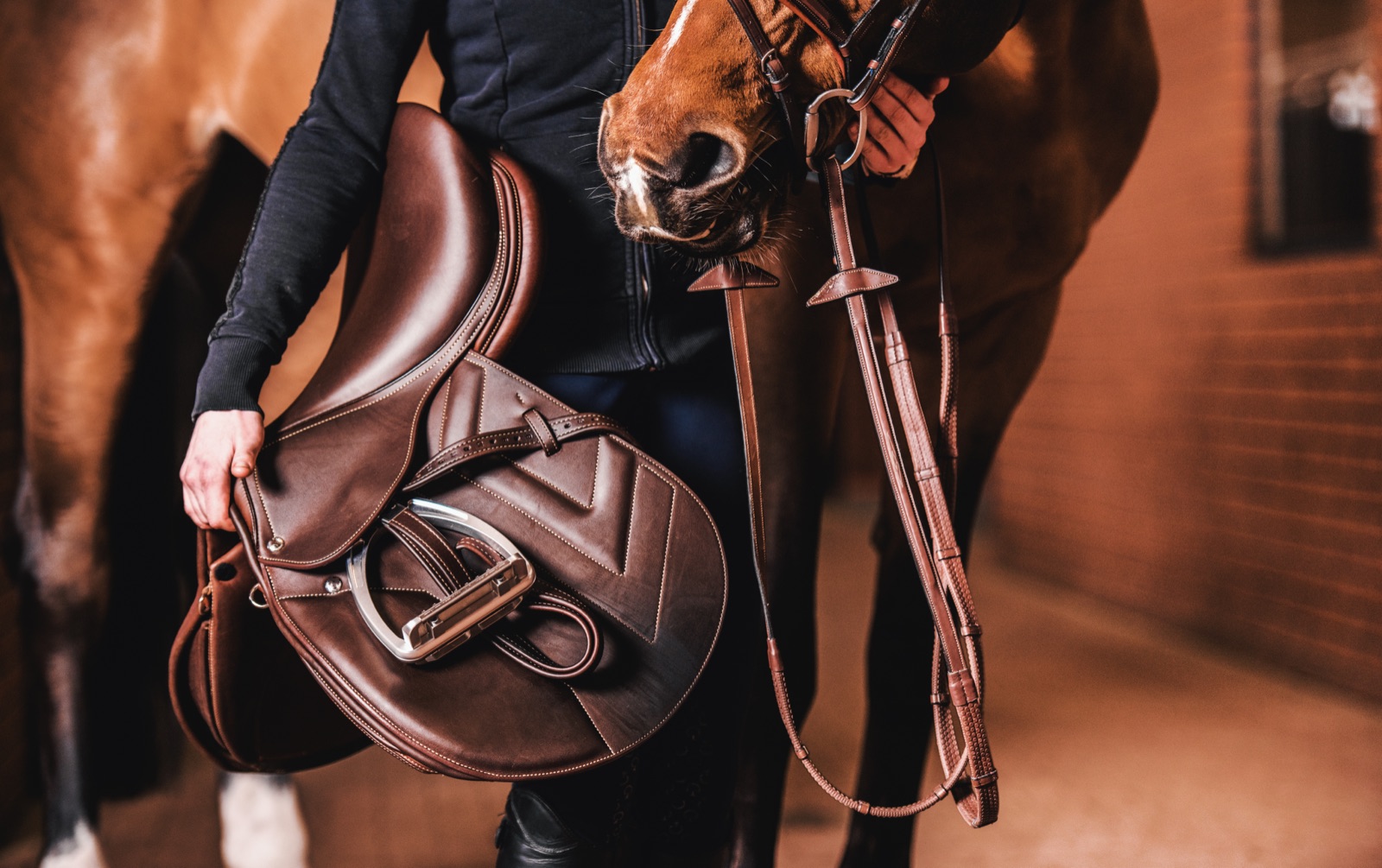 A rider holding a saddle and bridle beside their horse in the stable